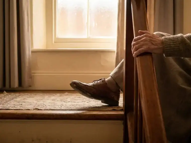 Versleten leren schoen die blijft haken op een ongelijke drempel, terwijl een oudere hand een houten trapleuning grijpt.