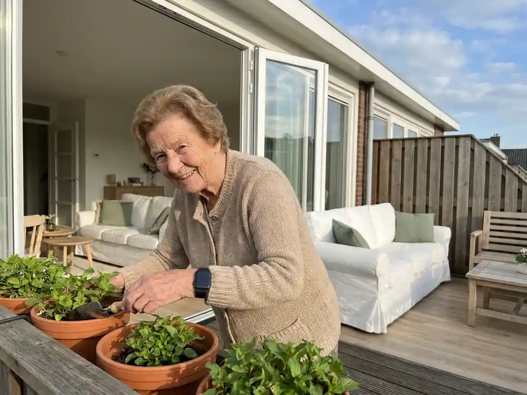 Oudere vrouw verzorgt balkonplanten met alarmhorloge om haar pols, lichte Nederlandse woning zichtbaar op de achtergrond.